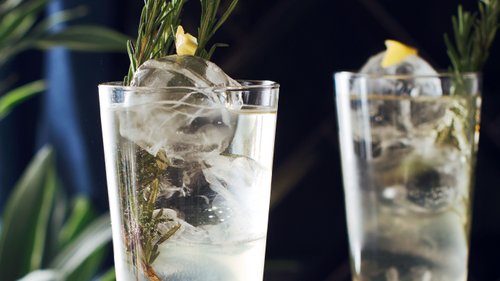 Zwei Gläser mit spritzigem Holunderblüten-Mocktail, verfeinert mit Rosmarinzweigen und Zitronenzeste, serviert auf einem eleganten Tablett