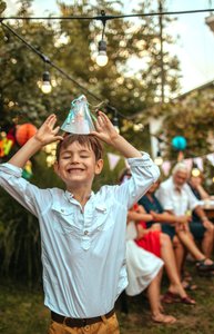 in glücklicher Junge feiert im Garten bei Tageslicht seinen Geburtstag, lacht und balanciert einen Partyhut auf seinem Kopf, während im Hintergrund Erwachsene an einem festlich dekorierten Tisch sitzen und den Moment genießen.