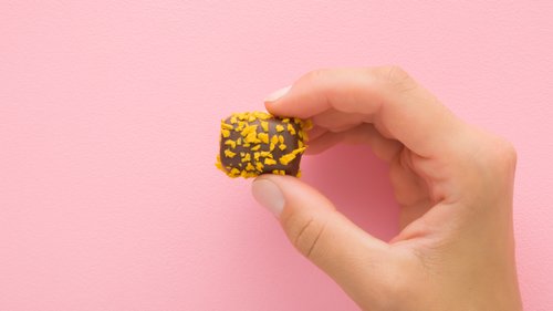 Eine Hand hält eine Praline vor einer rosa Wand, die mit goldgelben Splittern bestreut ist