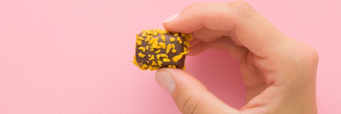 Eine Hand hält eine Praline vor einer rosa Wand, die mit goldgelben Splittern bestreut ist