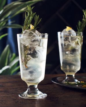 Zwei Gläser mit spritzigem Holunderblüten-Mocktail, verfeinert mit Rosmarinzweigen und Zitronenzeste, serviert auf einem eleganten Tablett