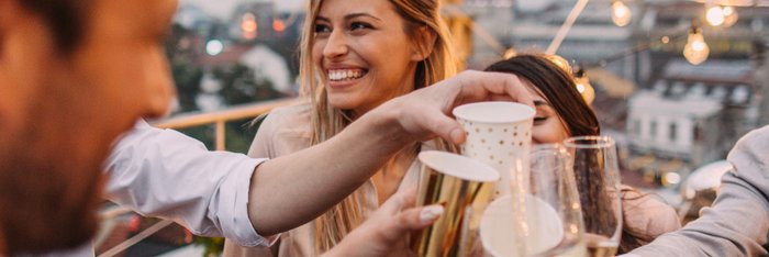 Fröhliche Gruppe von Freunden stößt mit "alkoholfreiem Sekt"* an, um einen gesunden und ausgelassenen Lebensstil auf einer Dachterrasse bei Dämmerung zu feiern.