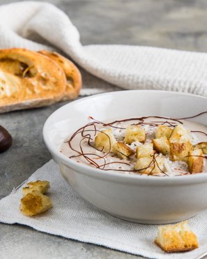 Eine Schale cremiger Maronensuppe, kunstvoll verziert, serviert mit Croutons und einem Hintergrund aus frischem Brot und einer Kastanie