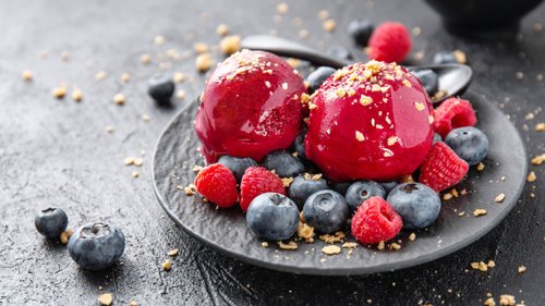 Zwei Kugeln leuchtend roter Himbeer-Granita auf einem dunklen Teller, garniert mit frischen Himbeeren und Blaubeeren, bestreut mit zerkrümelten Keksen