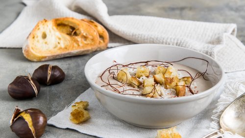 Eine Schale cremiger Maronensuppe, kunstvoll verziert, serviert mit Croutons und einem Hintergrund aus frischem Brot und einer Kastanie