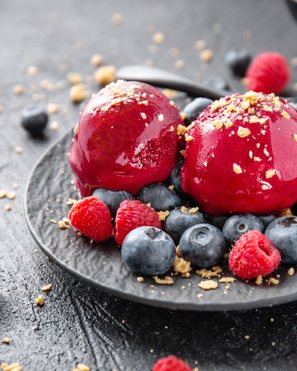 Zwei Kugeln leuchtend roter Himbeer-Granita auf einem dunklen Teller, garniert mit frischen Himbeeren und Blaubeeren, bestreut mit zerkrümelten Keksen