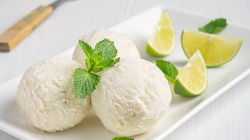 Drei Kugeln erfrischendes Sekt-Sorbet ohne Alkohol, serviert auf einer rechteckigen weißen Platte, garniert mit frischer Minze und Limettenspalten