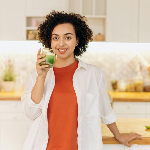 Lächelnde Frau mit lockigen Haaren hält ein Glas grünen Smoothie in der Küche